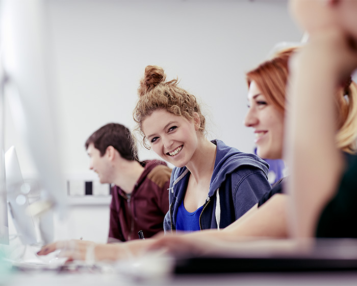 Student sat working at a desk, smiling, with other students on either side of her